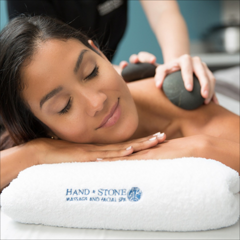 A woman with her eyes closed rests her head on her hands on top of a Hand & Stone towel while hands place stones on her back